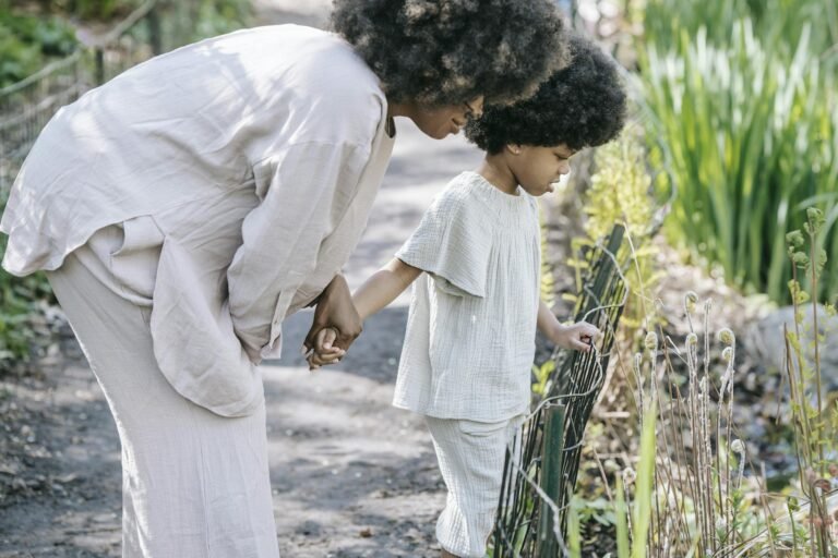 A woman and child with curly hair observe nature, holding hands by a garden path.